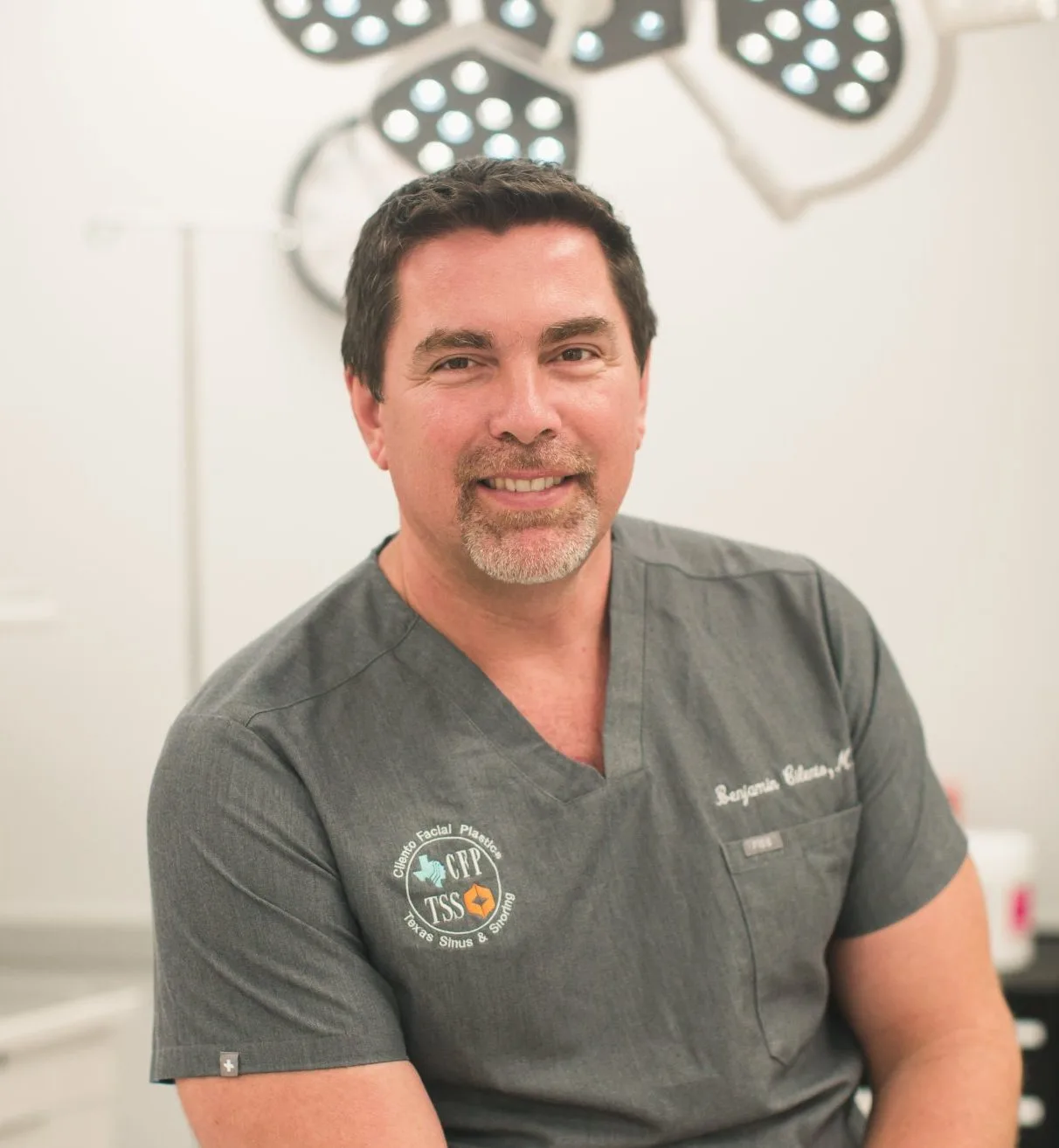 Dr. Ben Cilento smiling in surgical scrubs in an operating room with overhead surgical lights